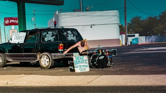 A person standing next to a black vehicle with signs advertising a deaf drummer. The person is reaching toward a drum kit set up at the side of the road. The setting appears to be a parking lot with a service station and signage in the background.