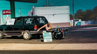 A person standing next to a black vehicle with signs advertising a deaf drummer. The person is reaching toward a drum kit set up at the side of the road. The setting appears to be a parking lot with a service station and signage in the background.