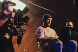Three individuals are engaged in a filmmaking or video production process inside a studio. One person operates a professional camera mounted on a rig, wearing a mask and cap. Two other people are seated, one intently looking at a laptop, creating an atmosphere of concentration and collaboration.