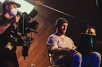 Three individuals are engaged in a filmmaking or video production process inside a studio. One person operates a professional camera mounted on a rig, wearing a mask and cap. Two other people are seated, one intently looking at a laptop, creating an atmosphere of concentration and collaboration.