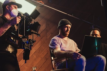 Three individuals are engaged in a filmmaking or video production process inside a studio. One person operates a professional camera mounted on a rig, wearing a mask and cap. Two other people are seated, one intently looking at a laptop, creating an atmosphere of concentration and collaboration.