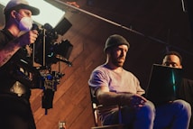 Three individuals are engaged in a filmmaking or video production process inside a studio. One person operates a professional camera mounted on a rig, wearing a mask and cap. Two other people are seated, one intently looking at a laptop, creating an atmosphere of concentration and collaboration.