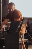 A person with a beard, wearing a brown shirt, is sitting on a director's chair working on a laptop. The setting appears to be a backstage area or a set with industrial elements visible in the background. The lighting casts warm tones, highlighting the person's concentration.
