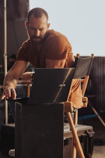 A person with a beard, wearing a brown shirt, is sitting on a director's chair working on a laptop. The setting appears to be a backstage area or a set with industrial elements visible in the background. The lighting casts warm tones, highlighting the person's concentration.