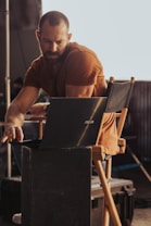 A person with a beard, wearing a brown shirt, is sitting on a director's chair working on a laptop. The setting appears to be a backstage area or a set with industrial elements visible in the background. The lighting casts warm tones, highlighting the person's concentration.