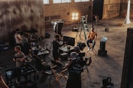 A group of people is gathered in an industrial-style setting, possibly a film or photography studio. Various equipment such as lights, cameras, tripods, and laptops can be seen in use. A person is seated on a director's chair while others appear to be setting up or adjusting the technical gear. The room has high ceilings with large windows and brick walls, creating a rustic atmosphere.