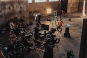 A group of people is gathered in an industrial-style setting, possibly a film or photography studio. Various equipment such as lights, cameras, tripods, and laptops can be seen in use. A person is seated on a director's chair while others appear to be setting up or adjusting the technical gear. The room has high ceilings with large windows and brick walls, creating a rustic atmosphere.