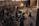 A group of people is gathered in an industrial-style setting, possibly a film or photography studio. Various equipment such as lights, cameras, tripods, and laptops can be seen in use. A person is seated on a director's chair while others appear to be setting up or adjusting the technical gear. The room has high ceilings with large windows and brick walls, creating a rustic atmosphere.