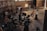 A group of people is gathered in an industrial-style setting, possibly a film or photography studio. Various equipment such as lights, cameras, tripods, and laptops can be seen in use. A person is seated on a director's chair while others appear to be setting up or adjusting the technical gear. The room has high ceilings with large windows and brick walls, creating a rustic atmosphere.