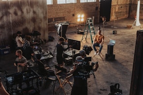 A group of people is gathered in an industrial-style setting, possibly a film or photography studio. Various equipment such as lights, cameras, tripods, and laptops can be seen in use. A person is seated on a director's chair while others appear to be setting up or adjusting the technical gear. The room has high ceilings with large windows and brick walls, creating a rustic atmosphere.