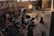 A group of people is gathered in an industrial-style setting, possibly a film or photography studio. Various equipment such as lights, cameras, tripods, and laptops can be seen in use. A person is seated on a director's chair while others appear to be setting up or adjusting the technical gear. The room has high ceilings with large windows and brick walls, creating a rustic atmosphere.