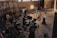A group of people is gathered in an industrial-style setting, possibly a film or photography studio. Various equipment such as lights, cameras, tripods, and laptops can be seen in use. A person is seated on a director's chair while others appear to be setting up or adjusting the technical gear. The room has high ceilings with large windows and brick walls, creating a rustic atmosphere.