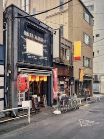 A narrow street scene showcases a cozy Japanese eatery with signs in Japanese script. The entrance is adorned with warm, inviting lanterns and an array of signs and menus displayed outside. Two bicycles are parked nearby, and the surrounding buildings are tall with a somewhat urban industrial aesthetic.