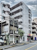 A juxtaposition of two buildings with contrasting architectural styles. An old, weathered structure with visible signs of decay stands adjacent to a modern, multi-story apartment building. The newer building features sleek lines and balconies. A small convenience store is integrated into the modern building's ground level. Power lines crisscross overhead, and a parked truck is visible on the street beside the older building.