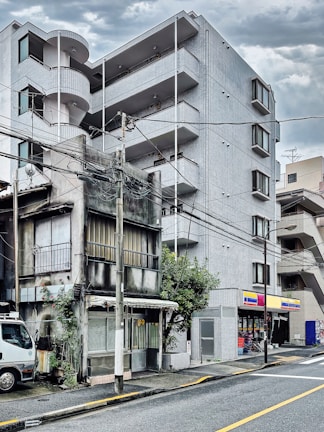 A juxtaposition of two buildings with contrasting architectural styles. An old, weathered structure with visible signs of decay stands adjacent to a modern, multi-story apartment building. The newer building features sleek lines and balconies. A small convenience store is integrated into the modern building's ground level. Power lines crisscross overhead, and a parked truck is visible on the street beside the older building.