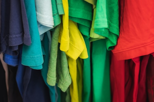 A collection of colorful t-shirts hanging closely together, showcasing an array of vibrant colors including shades of blue, green, yellow, and red. The shirts appear to be made of soft fabric and are displayed side by side, giving a sense of variety and brightness.