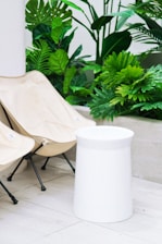 A minimalist outdoor setup featuring two beige folding chairs and a cylindrical white stool on a light tiled floor. Lush green tropical plants in the background add a fresh, natural element to the scene.