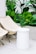 A minimalist outdoor setup featuring two beige folding chairs and a cylindrical white stool on a light tiled floor. Lush green tropical plants in the background add a fresh, natural element to the scene.