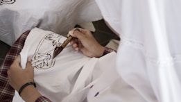 A person is holding a canting tool and applying wax to a piece of fabric as part of a batik-making process. The fabric is white with a brown design being traced, and the person wears plaid clothing. The scene suggests a focus on traditional textile craftsmanship.