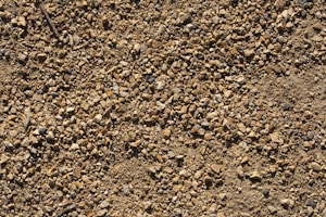 A close-up view of a ground surface covered with small, angular gravel and dirt. The stones vary in size and color, with shades ranging from light tan to darker browns and some grayish tones. Sparse twigs and organic debris are scattered among the gravel.