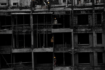 A construction site featuring a multi-story building in progress. Several workers wearing yellow helmets are scattered across different floors, with some engaged in activities such as carrying materials or working on the structure. The building appears to be made of concrete, and there are numerous steel rods protruding from the top floor.