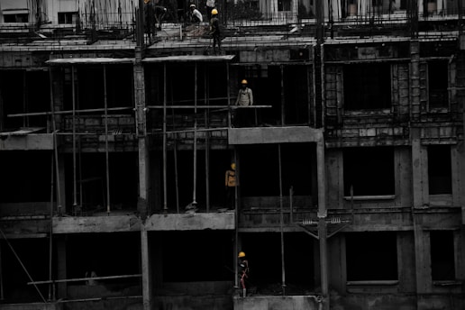A construction site featuring a multi-story building in progress. Several workers wearing yellow helmets are scattered across different floors, with some engaged in activities such as carrying materials or working on the structure. The building appears to be made of concrete, and there are numerous steel rods protruding from the top floor.