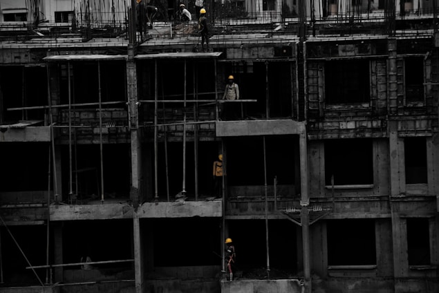 A construction site featuring a multi-story building in progress. Several workers wearing yellow helmets are scattered across different floors, with some engaged in activities such as carrying materials or working on the structure. The building appears to be made of concrete, and there are numerous steel rods protruding from the top floor.