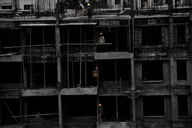 A construction site featuring a multi-story building in progress. Several workers wearing yellow helmets are scattered across different floors, with some engaged in activities such as carrying materials or working on the structure. The building appears to be made of concrete, and there are numerous steel rods protruding from the top floor.