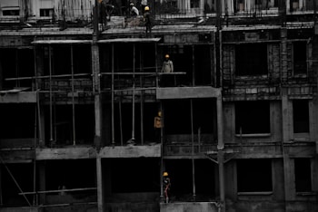 A construction site featuring a multi-story building in progress. Several workers wearing yellow helmets are scattered across different floors, with some engaged in activities such as carrying materials or working on the structure. The building appears to be made of concrete, and there are numerous steel rods protruding from the top floor.