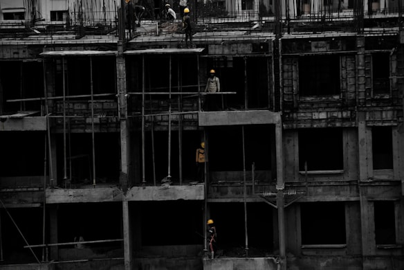A construction site featuring a multi-story building in progress. Several workers wearing yellow helmets are scattered across different floors, with some engaged in activities such as carrying materials or working on the structure. The building appears to be made of concrete, and there are numerous steel rods protruding from the top floor.