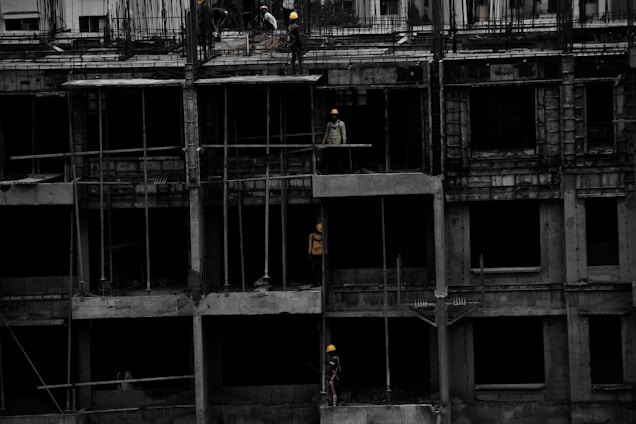 A construction site featuring a multi-story building in progress. Several workers wearing yellow helmets are scattered across different floors, with some engaged in activities such as carrying materials or working on the structure. The building appears to be made of concrete, and there are numerous steel rods protruding from the top floor.