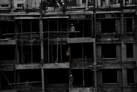 A construction site featuring a multi-story building in progress. Several workers wearing yellow helmets are scattered across different floors, with some engaged in activities such as carrying materials or working on the structure. The building appears to be made of concrete, and there are numerous steel rods protruding from the top floor.