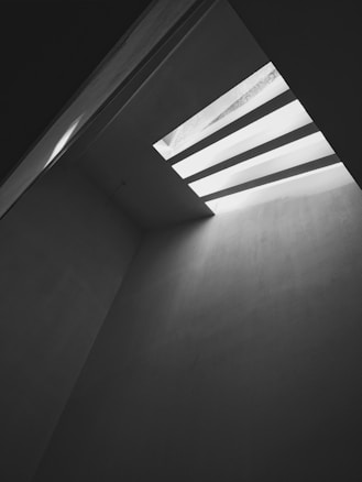 A stark, minimalistic architectural space with a high ceiling. Light streams in from a rectangular skylight composed of parallel slats, casting sharp shadows on the walls. The space feels open yet confined, with smooth, bare surfaces highlighted by contrasting light and dark areas.