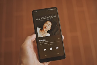 A hand holding a smartphone that displays a music player app. The screen shows an album cover with a person lying down, and text indicating the song and artist name 'My Little Airport' in both English and Chinese. The background is a wooden floor with a warm brown tone.