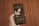 A hand holding a smartphone that displays a music player app. The screen shows an album cover with a person lying down, and text indicating the song and artist name 'My Little Airport' in both English and Chinese. The background is a wooden floor with a warm brown tone.