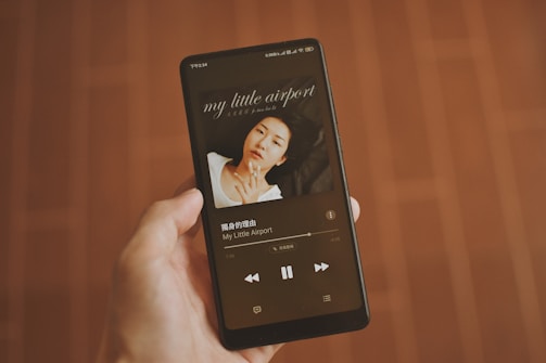 A hand holding a smartphone that displays a music player app. The screen shows an album cover with a person lying down, and text indicating the song and artist name 'My Little Airport' in both English and Chinese. The background is a wooden floor with a warm brown tone.