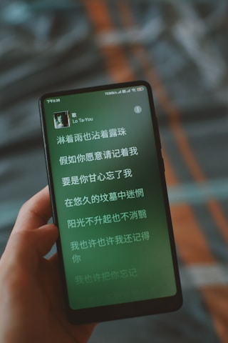A smartphone displaying song lyrics in Chinese on its screen. The phone is being held in a person's hand, and the background is blurred, featuring soft colors and patterns.