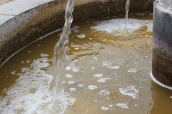 Water flows from two spouts into a circular basin filled with murky, brownish water that has foam and bubbles on the surface. The basin appears concrete and shows signs of wear and dirt.