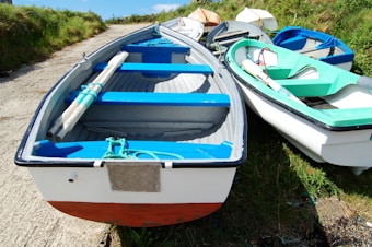 Several small boats are resting on a grassy and gravel area near a path, with vibrant blue and green interiors. The scene is outdoors, with green grass surrounding the boats and a clear blue sky above.