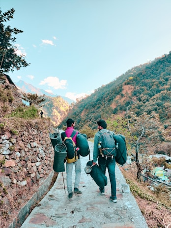 Two people are walking on a stone-paved path in a mountainous area, carrying backpacks and rolled-up sleeping mats. The background features a hillside covered with trees and sparse vegetation. There is a clear sky with a few clouds and some structures and houses visible in the distance.