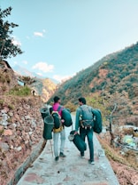 Two people are walking on a stone-paved path in a mountainous area, carrying backpacks and rolled-up sleeping mats. The background features a hillside covered with trees and sparse vegetation. There is a clear sky with a few clouds and some structures and houses visible in the distance.