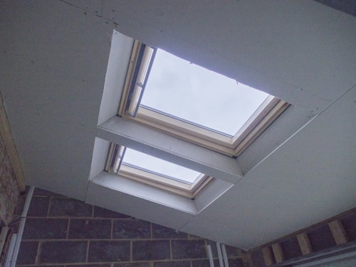 A view of two large, rectangular skylights set into a sloped ceiling, allowing natural light to enter a room. The ceiling features unfinished drywall and visible bricks on one wall corner.
