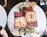 Two rectangular pizzas topped with cheese, tomato slices, and herbs are placed on a black rectangular slate. Next to the pizzas are small bowls with red dipping sauces. Four beverages garnished with mint leaves and straws are set on the same table.