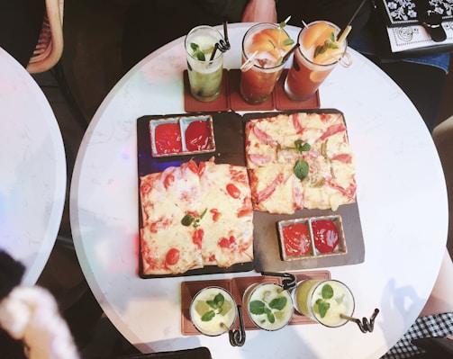 Two rectangular pizzas topped with cheese, tomato slices, and herbs are placed on a black rectangular slate. Next to the pizzas are small bowls with red dipping sauces. Four beverages garnished with mint leaves and straws are set on the same table.