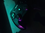 A black gaming controller is illuminated by a mix of teal and purple lighting, highlighting its buttons and thumbsticks. The logo and select button are visible, creating a sleek and modern appearance.
