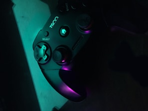 A black gaming controller is illuminated by a mix of teal and purple lighting, highlighting its buttons and thumbsticks. The logo and select button are visible, creating a sleek and modern appearance.