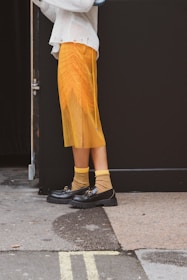 An individual stands outdoors wearing a flowy, pleated yellow skirt, yellow and black striped socks, and black loafers. The person is also wearing a white long-sleeved knit sweater. The background includes a dark wall and an entryway.