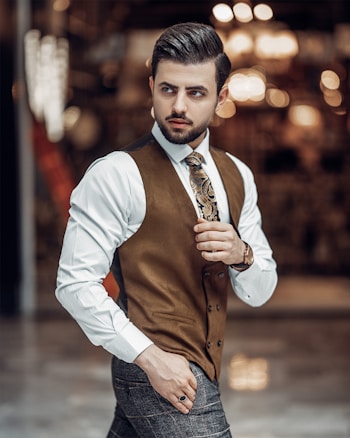 A well-dressed man is seen wearing a white shirt, patterned tie, and a brown vest with a watch on his wrist. He is in a stylish, modern setting with bokeh lights in the background, giving the image a sophisticated and elegant atmosphere.