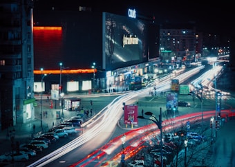 A bustling city intersection at night with bright light trails from moving vehicles. The streets are lined with various buildings and an illuminated billboard. Numerous streetlights and neon signs add to the vibrant atmosphere. People are walking along sidewalks, and parked cars line one side of the street.