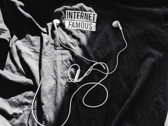A black t-shirt is laid out with the words 'Internet Famous' printed on it. White earbuds with tangled cords rest on top of the shirt, creating a contrast against the dark background.